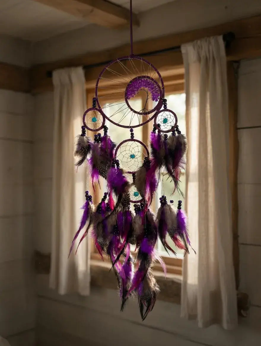 A purple dreamcatcher hanging in front of a window with white curtains. The dreamcatcher features multiple hanging rings, feathers in shades of purple, brown, and black, and small turquoise bead details.
