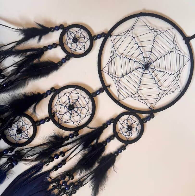 A black dreamcatcher wall hanging with four circular hoops, woven web patterns, dark beads, and black feathers, photographed against a plain light beige background.