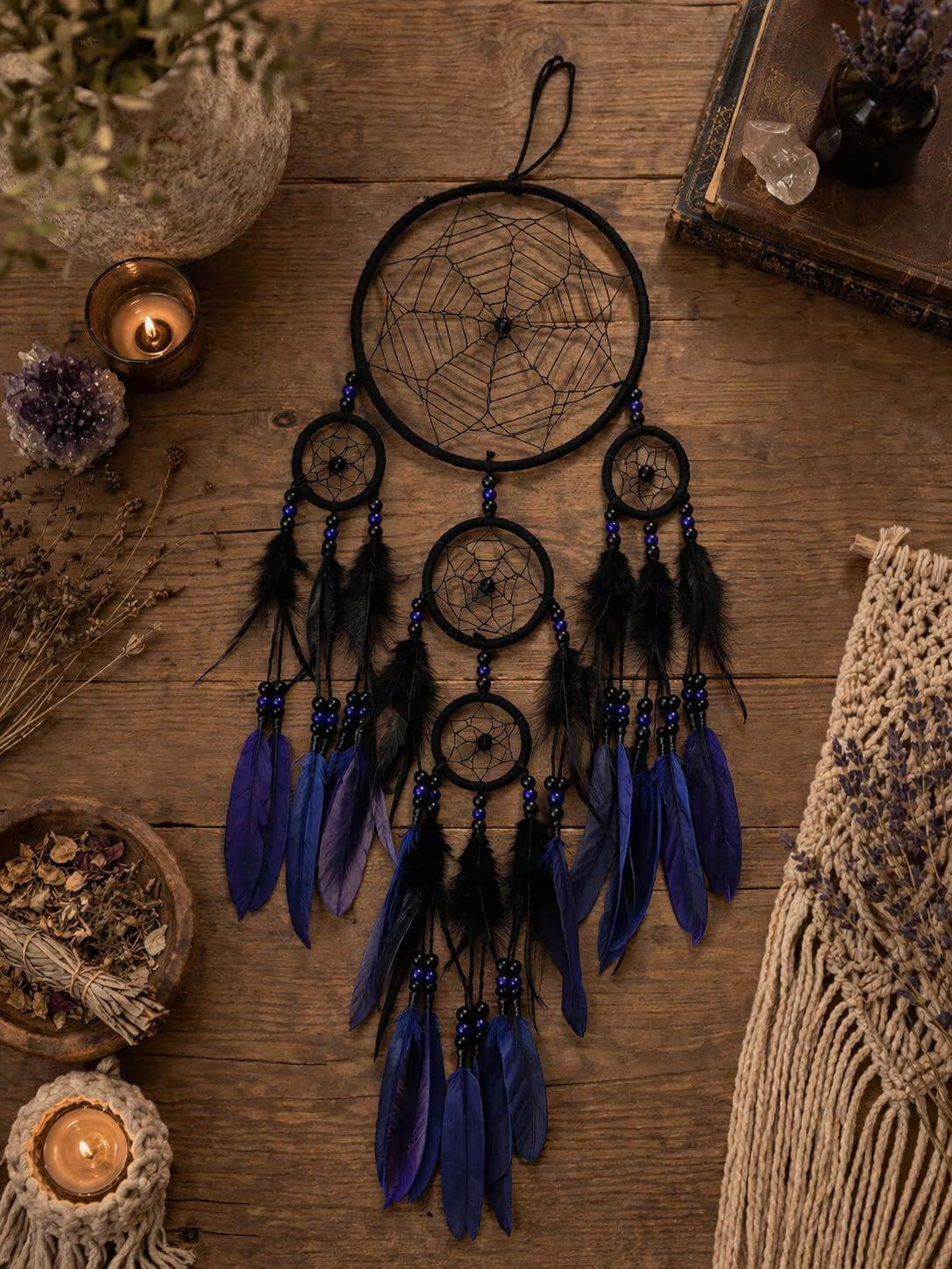 A bohemian dreamcatcher with three hanging rings, black webbing, and dark blue and black feathers, displayed on a rustic wooden table surrounded by candles, dried flowers, and a macrame textile.