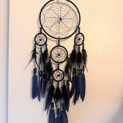 A large black dreamcatcher with multiple hanging rings, dark blue feathers, and black beads, displayed against a plain off-white wall.