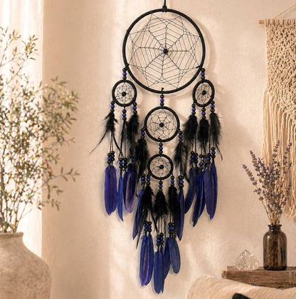 A black circular dreamcatcher wall hanging with multiple webbed rings, black feathers, and long blue feathers, displayed against a beige wall in a styled interior setting.