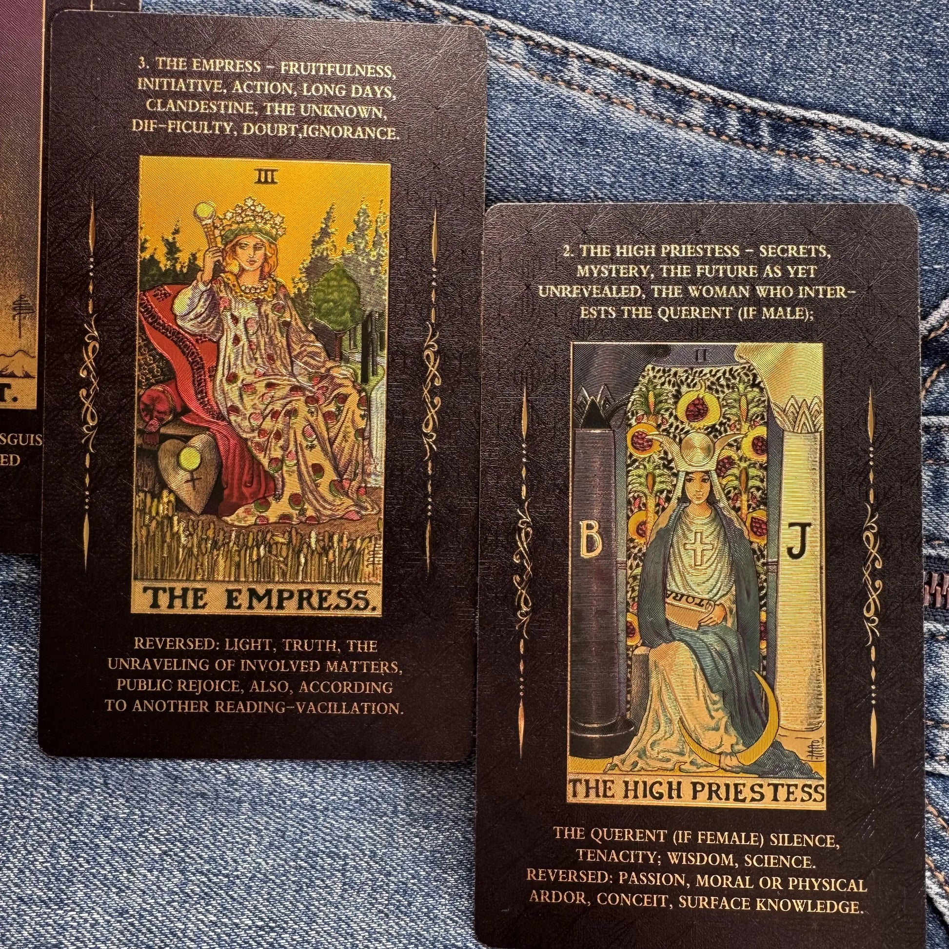 Two tarot cards, 'The Empress' and 'The High Priestess', displayed on a blue denim fabric background. The cards feature dark brown borders with gold text and detailed gold and red illustrations of the figures.