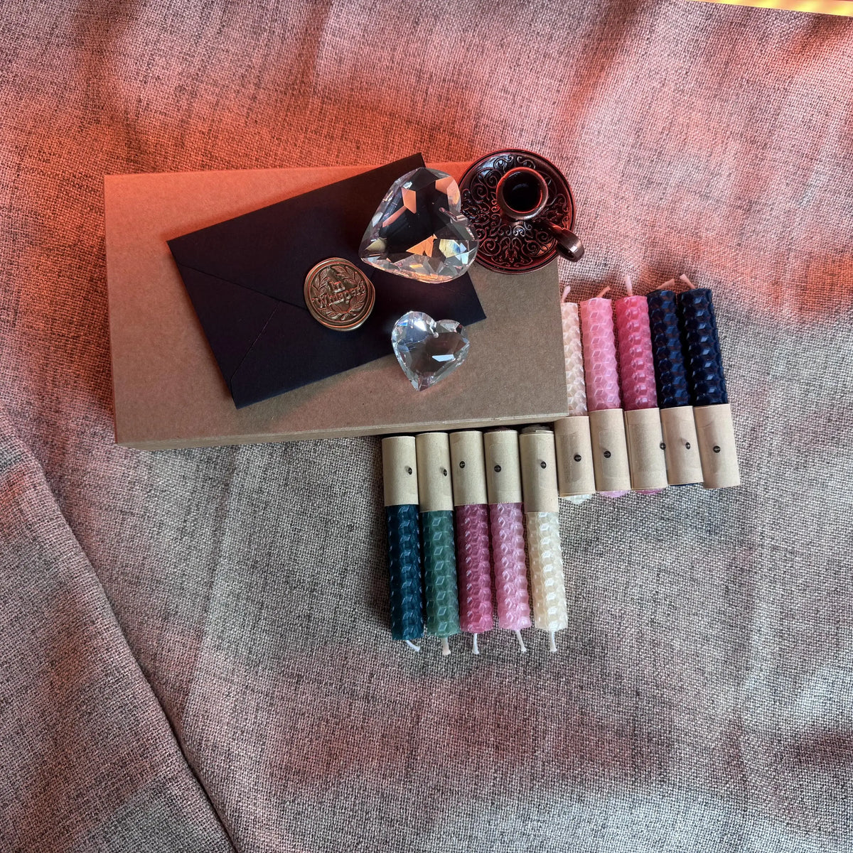A styled flat-lay of natural beeswax candles melts in various colors (pink, blue, teal, cream) with wooden wicks, arranged next to a kraft paper envelope with a wax seal and decorative props on a textured gray fabric background.