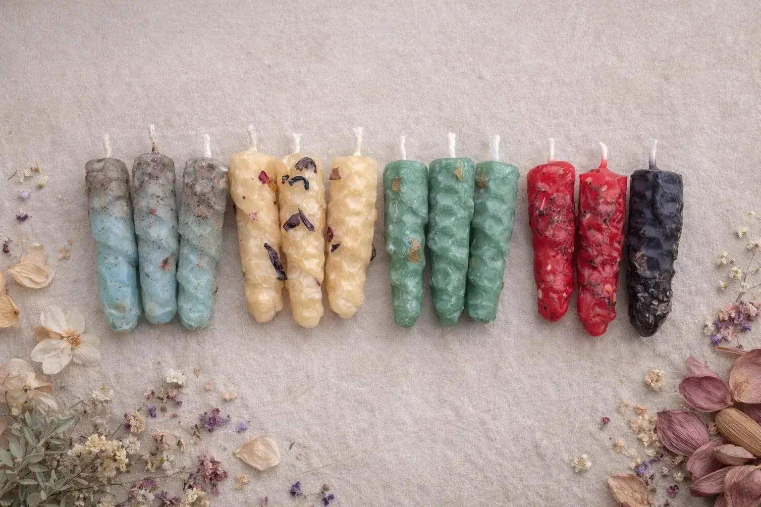 Row of colorful  natural beeswax candles on a textured surface with dried flowers and herbs.