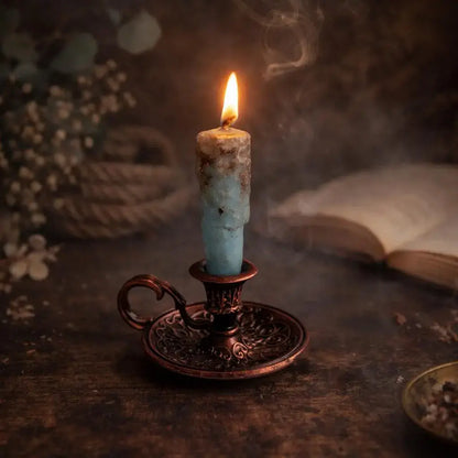 A lit blue  natural beeswax candle in an ornate dark bronze holder on a rustic wooden surface, with a blurred open book and dried flowers in the background.