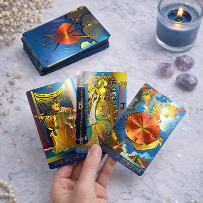 A hand holds three tarot cards from an oracle card deck, with a stack of the same deck and a blue candle on a light marble surface. The cards feature colorful illustrated figures and symbols, and the scene includes crystals and glitter.