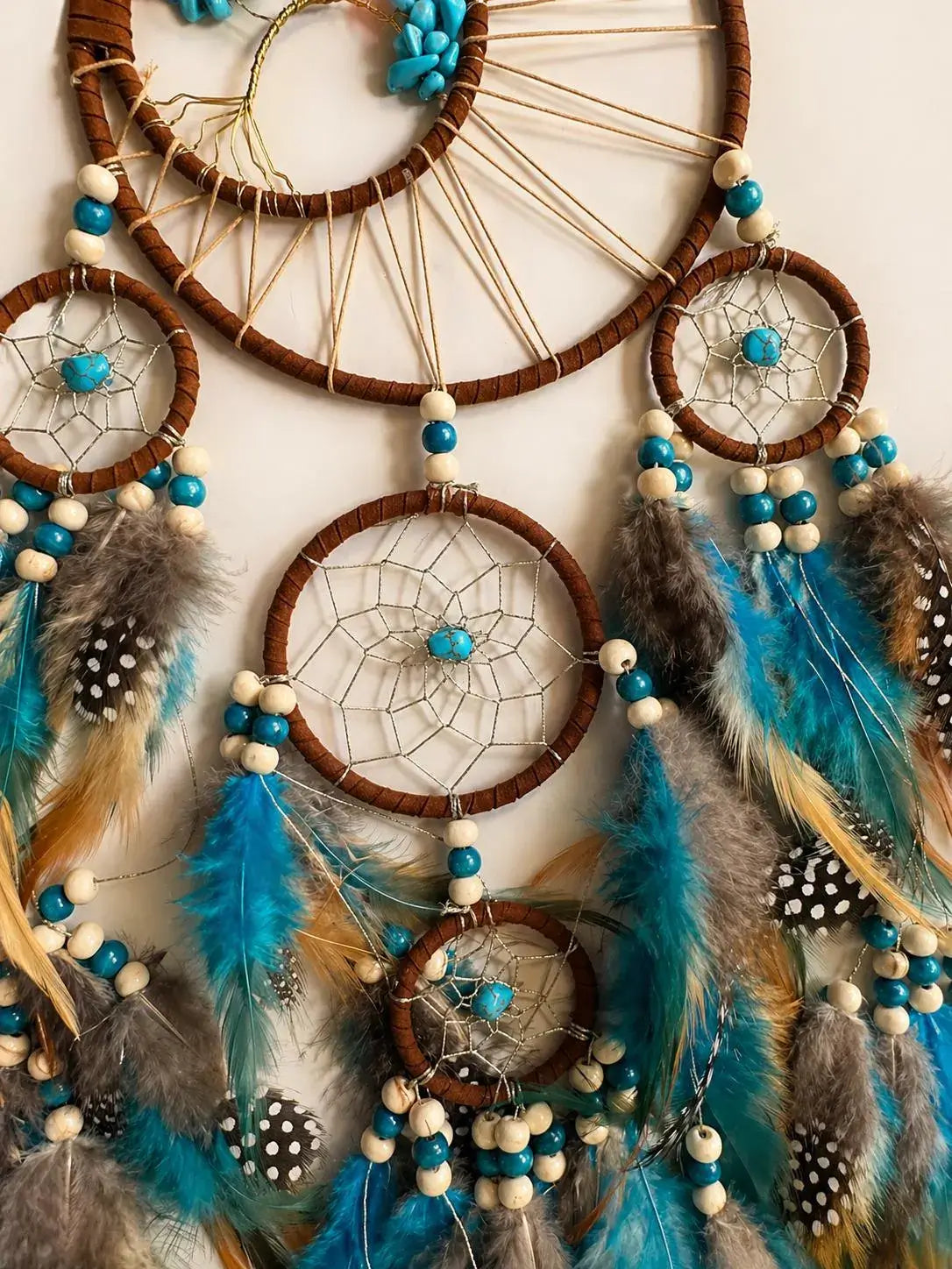 A large dreamcatcher wall hanging with multiple woven hoops, turquoise beads, white wooden beads, brown feathers, and blue, teal, gray, and tan feathers against a plain light background.