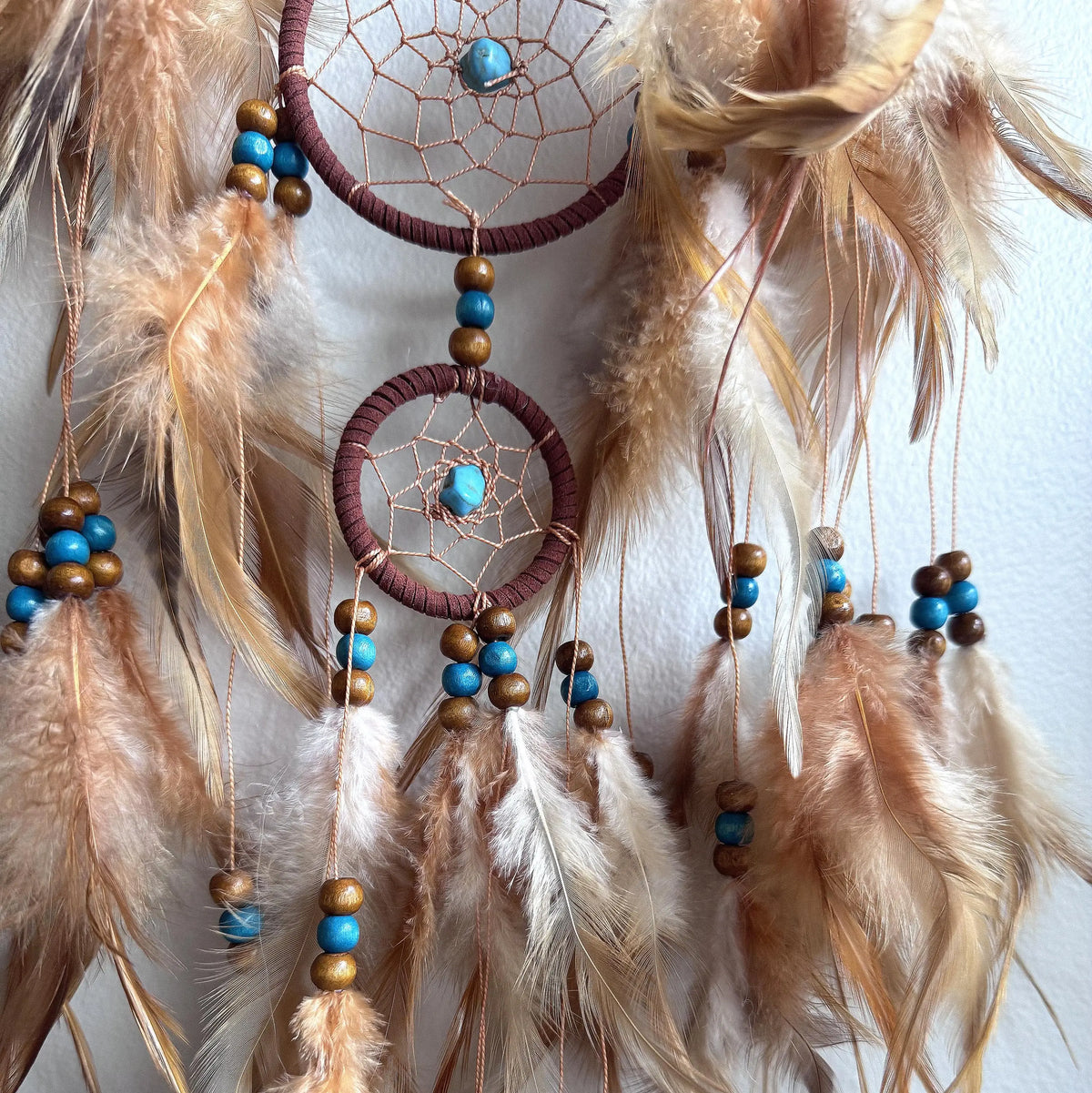A bohemian dreamcatcher wall hanging with two circular webbed centers, tan and brown feathers, turquoise blue beads, and wooden beads, shown against a plain white wall.