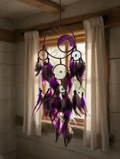 A purple dreamcatcher hanging in front of a window with white curtains. The dreamcatcher features multiple hanging rings, feathers in shades of purple, brown, and black, and small turquoise bead details.