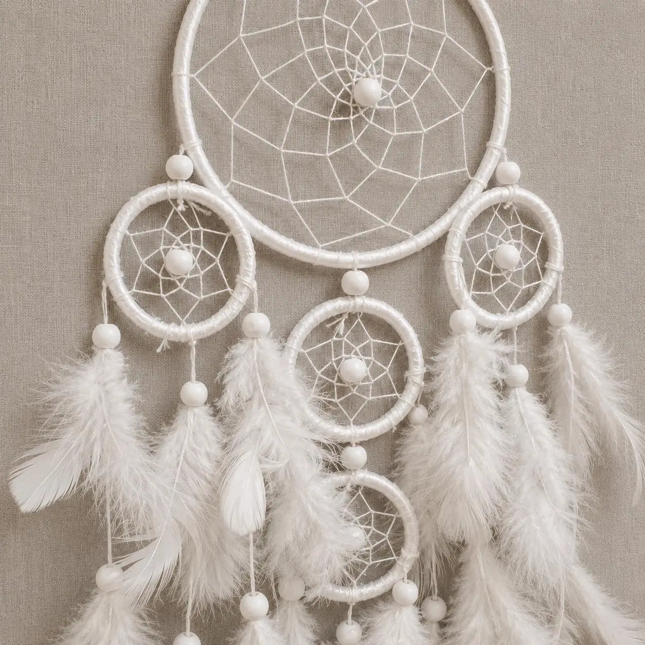 A white dreamcatcher with multiple woven rings, white beads, and soft white feathers hanging against a neutral beige textured background.
