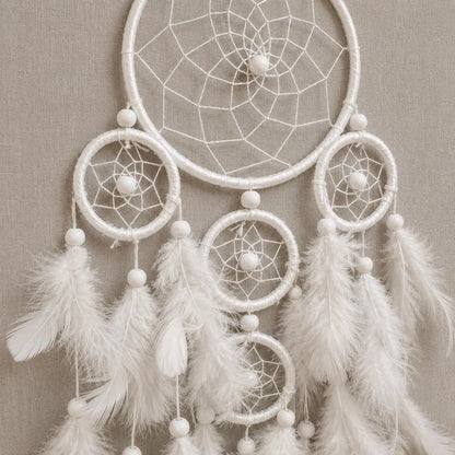 A white dreamcatcher with multiple woven rings, white beads, and soft white feathers hanging against a neutral beige textured background.