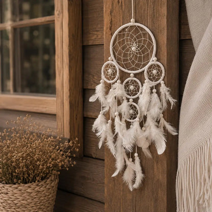 A white bohemian dreamcatcher with multiple woven rings, feathers, and beads hanging on a rustic wooden door next to a woven basket of dried flowers.