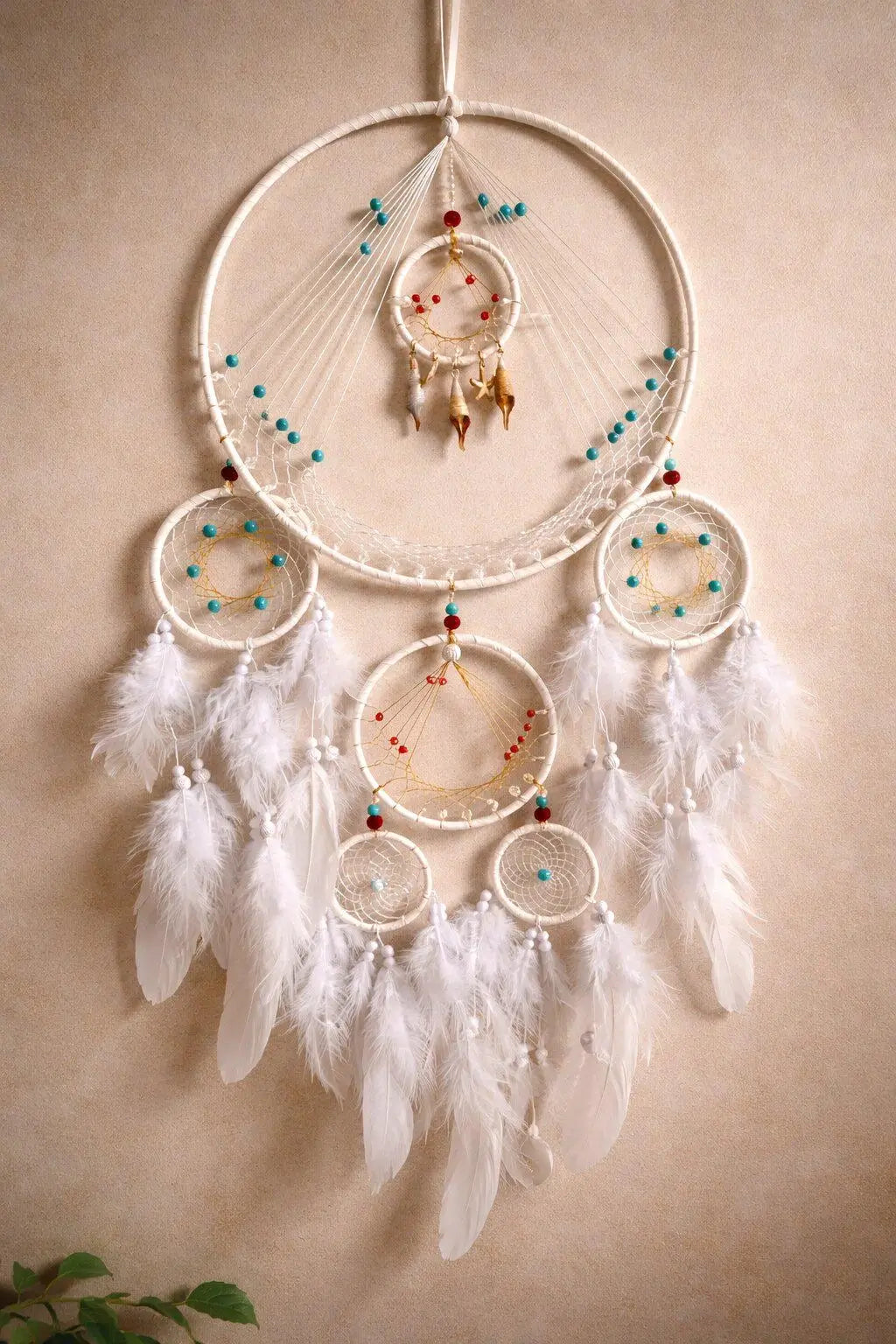 A large bohemian dreamcatcher wall hanging with multiple white feathers, white webbed hoops, and turquoise and red bead accents, hanging against a warm beige wall.