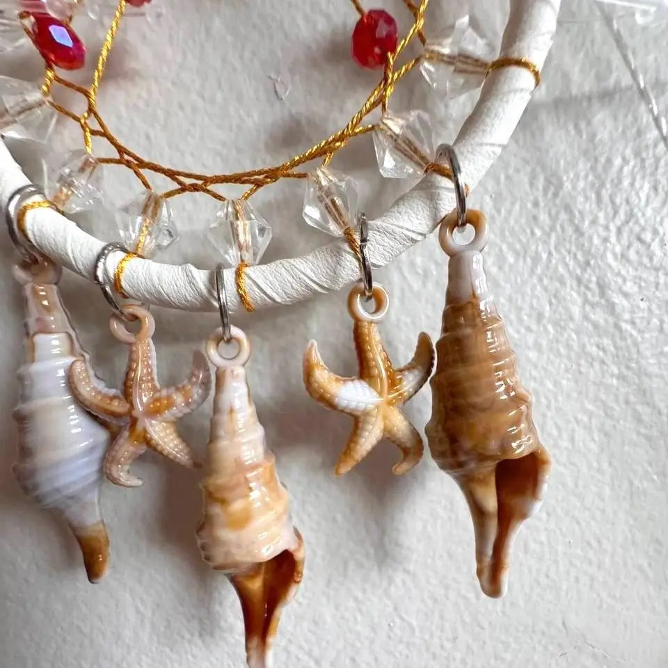 A close-up of a jewelry display featuring several seashell and starfish charms hanging from a white wrapped display rod with gold wire and clear faceted beads.