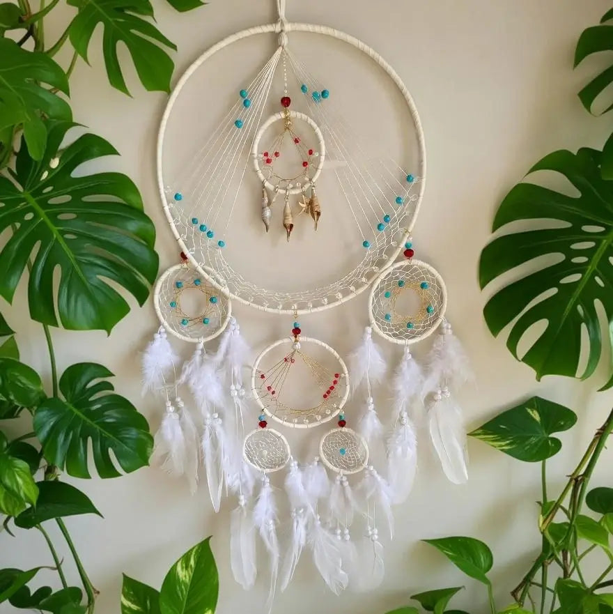 A large white dreamcatcher wall hanging with multiple concentric hoops, white feathers, and small turquoise and red bead accents, displayed against a light wall with green monstera leaves around it.