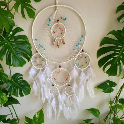 A large white dreamcatcher wall hanging with multiple concentric hoops, white feathers, and small turquoise and red bead accents, displayed against a light wall with green monstera leaves around it.