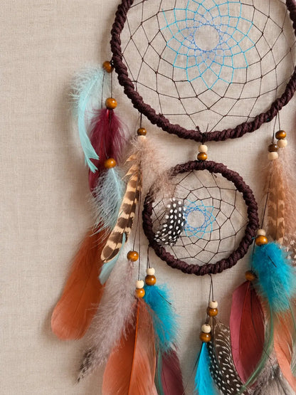 A bohemian dreamcatcher wall hanging with two woven dark brown hoops, colorful feathers in turquoise, rust orange, pink, and brown, and wooden beads, displayed against a neutral beige linen background.