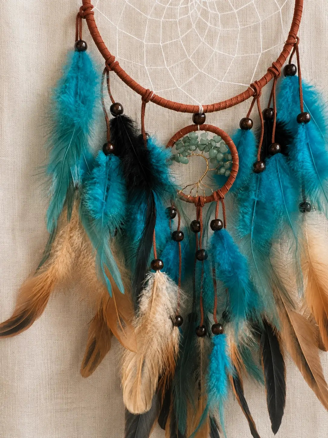 A dreamcatcher with turquoise, black, and tan feathers, brown wrapped cords, and dark wooden beads, hanging against a light beige linen background.