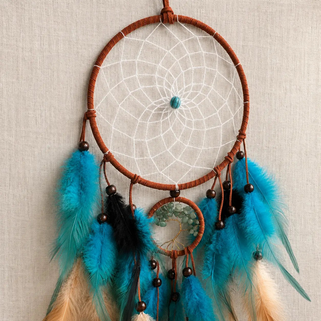 A dreamcatcher with a brown woven hoop, white webbing, turquoise blue feathers, black beads, and feathers in turquoise, black, and cream, hanging against a textured beige fabric background.