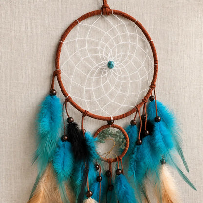A dreamcatcher with a brown woven hoop, white webbing, turquoise blue feathers, black beads, and feathers in turquoise, black, and cream, hanging against a textured beige fabric background.