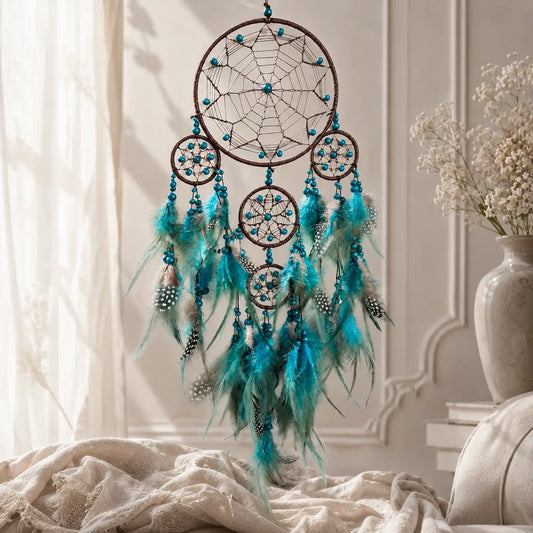 A dreamcatcher wall hanging with multiple brown hoops, turquoise beads, and long teal feathers, displayed in a softly lit room with draped fabric and a vase of flowers in the background.